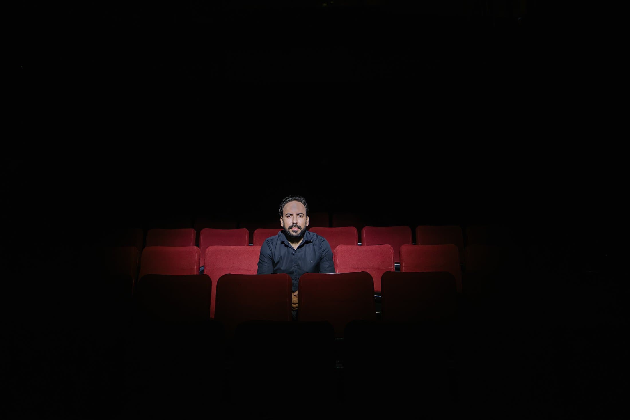 Homem sentado sozinho em uma plateia de teatro, iluminado em meio às poltronas vazias